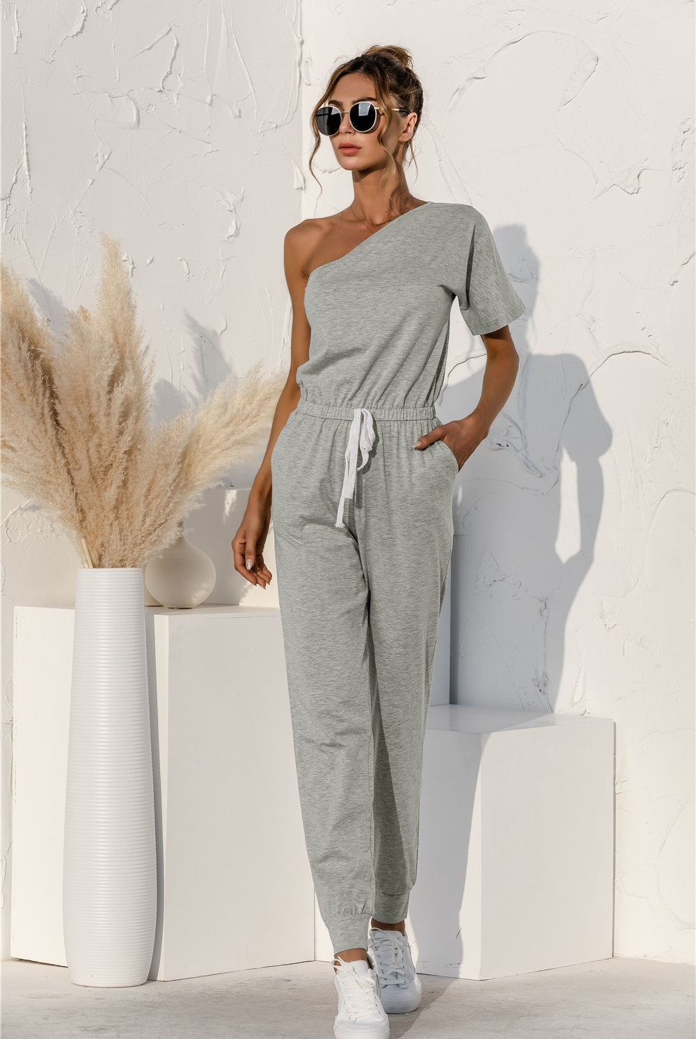 A model poses confidently in a one-shoulder jumpsuit with a drawstring waist, paired with classic white sneakers, ready for a relaxed yet fashionable day out.