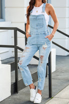 A woman poses confidently in light wash distressed denim overalls, paired with a simple white tank top and white sneakers for an effortlessly cool urban look.