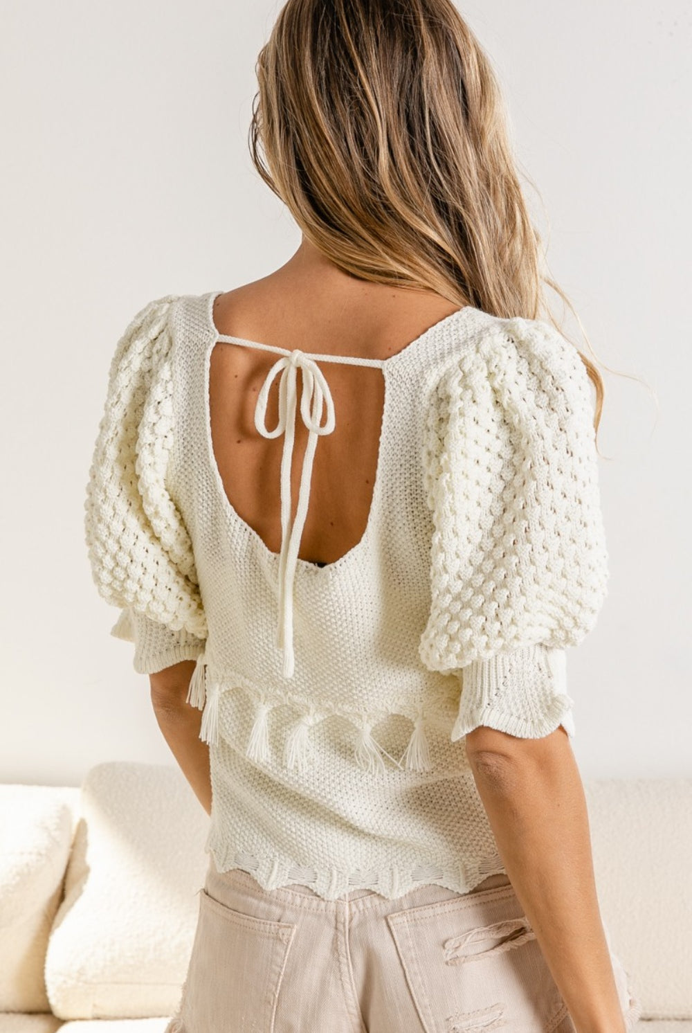 A woman models a textured tassel sweater with puff sleeves and a round neckline, styled with frayed light beige shorts for a cozy yet chic look.