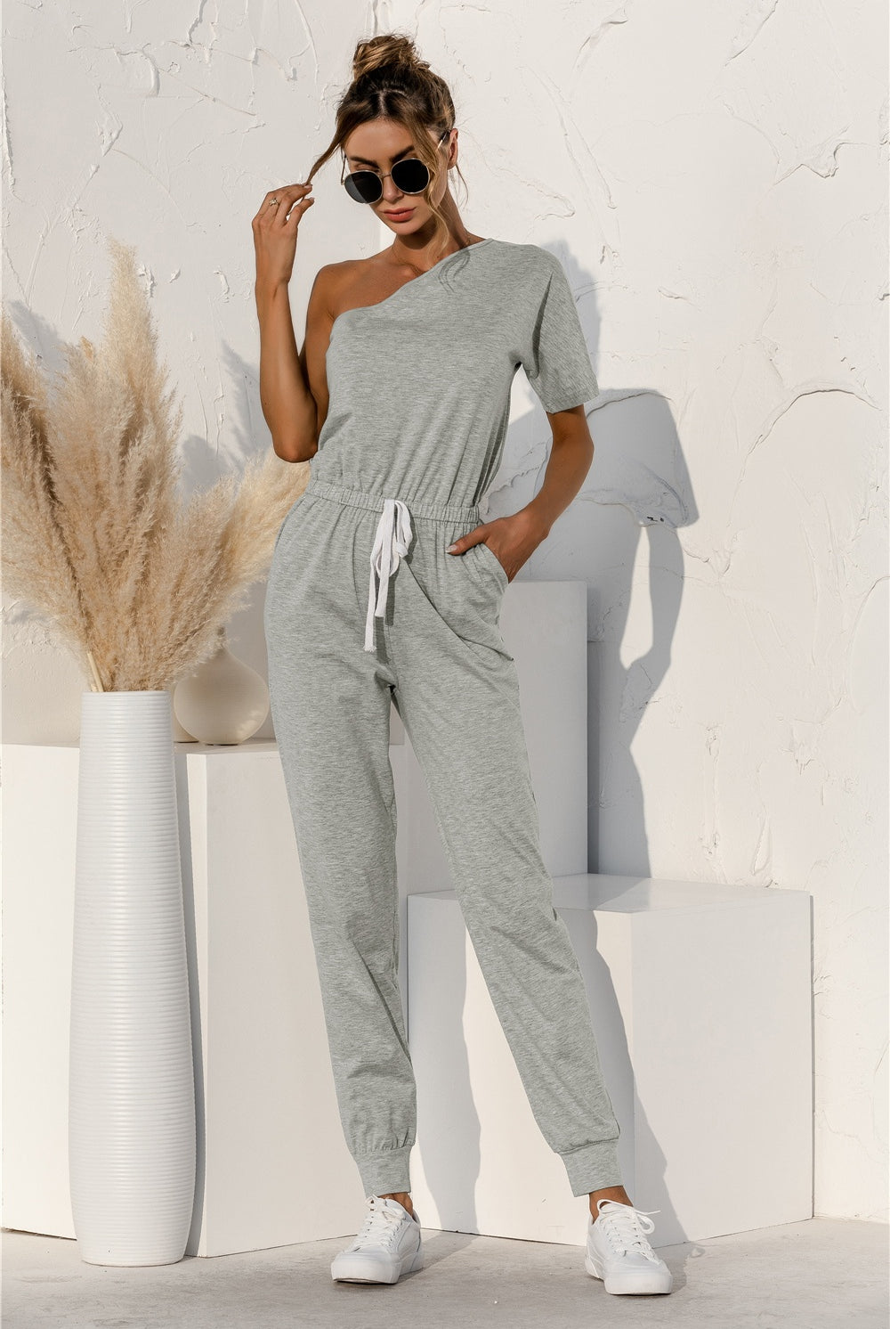 A model poses confidently in a one-shoulder jumpsuit with a drawstring waist, paired with classic white sneakers, ready for a relaxed yet fashionable day out.