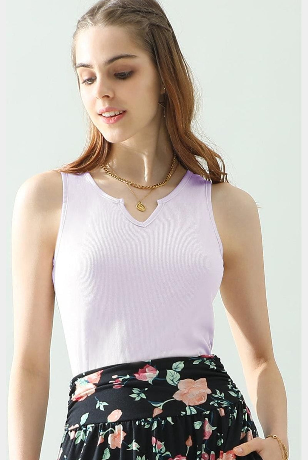 A model beams in a sleeveless knit tank top featuring a subtle ribbed texture and a scoop neckline, styled with a floral-print skirt. She is accessorized with layered necklaces and bracelets, presenting a chic and approachable look. The tank's knit design offers a cozy yet elegant element, available in a range of colors to suit different styles and preferences.