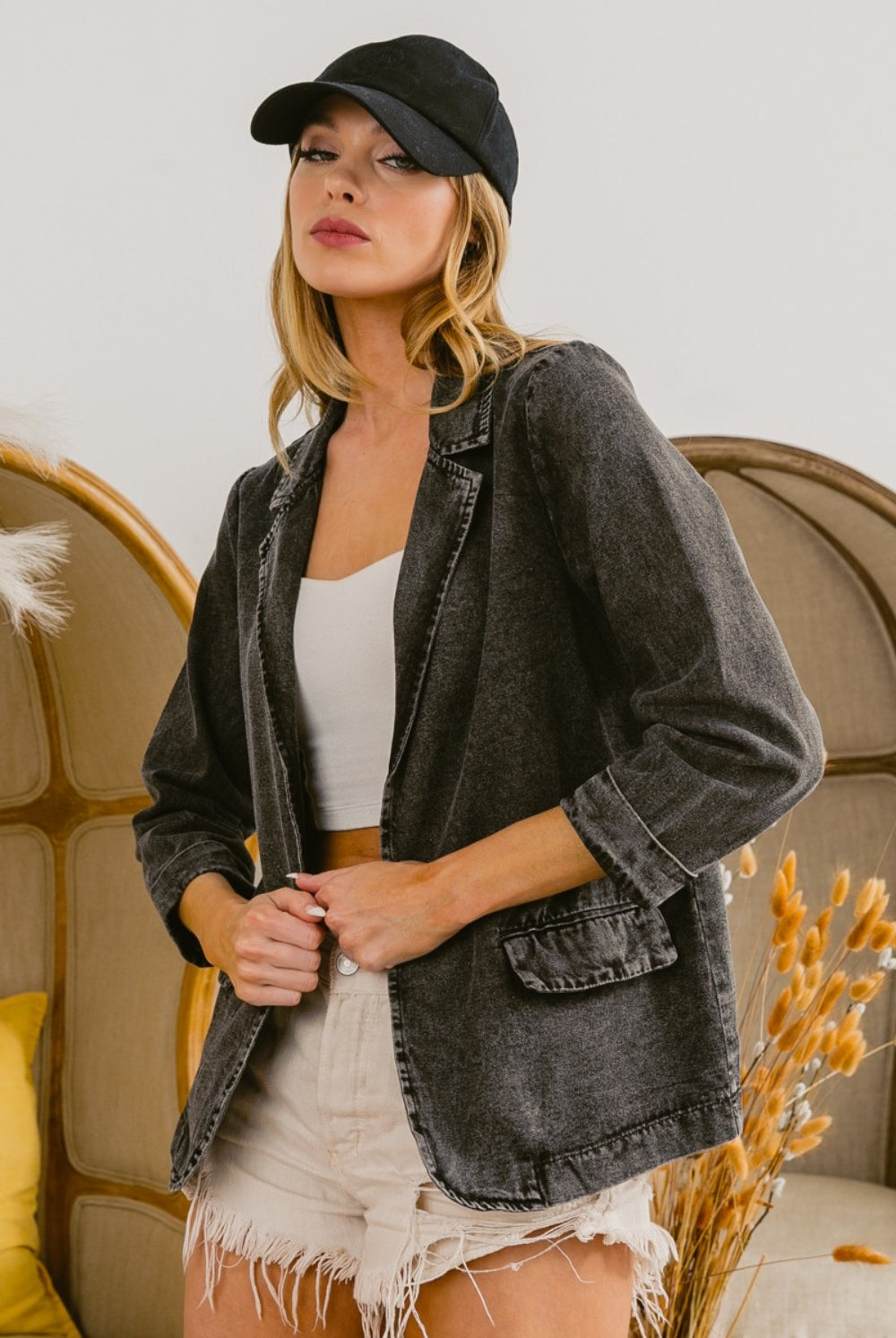 A woman wearing a chic denim blazer with a textured finish, styled with a simple white top, distressed cream shorts, and accessorized with a black cap for a smart-casual look.