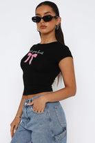 A woman wearing a fitted, crew-neck graphic t-shirt with the phrase "Protect your heart" above a ribbon design, paired with casual denim jeans and accessorized with chic sunglasses and simple jewelry. The shirt has a cropped cut, offering a modern and stylish look.