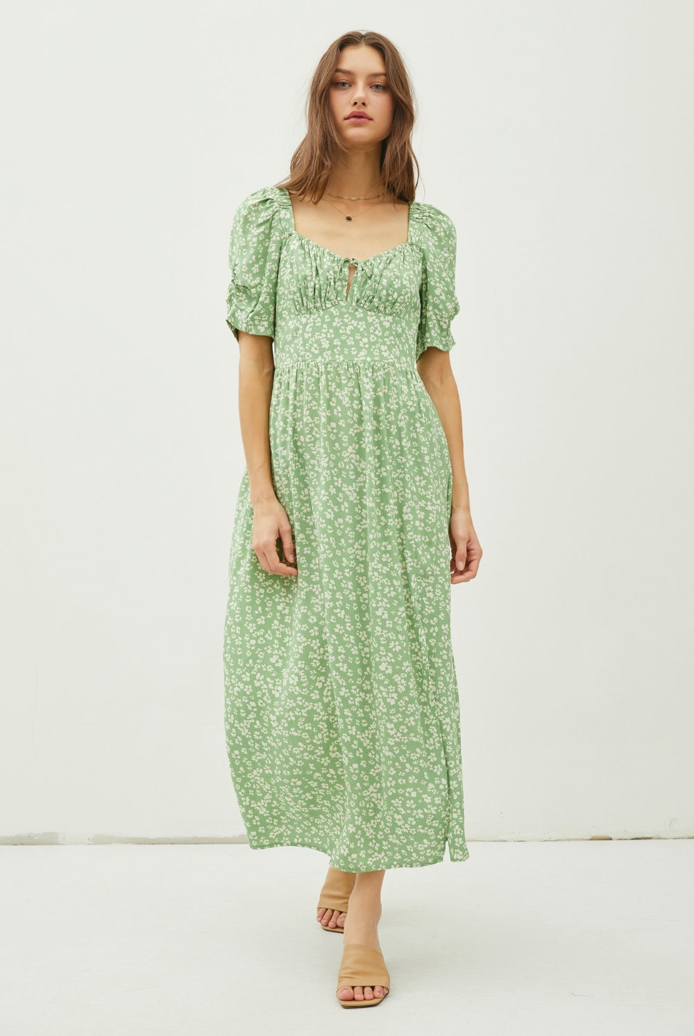 A poised woman exhibits a serene and romantic look in a sage green maxi dress with a delicate floral print and a tasteful back slit, perfect for spring and summer occasions.