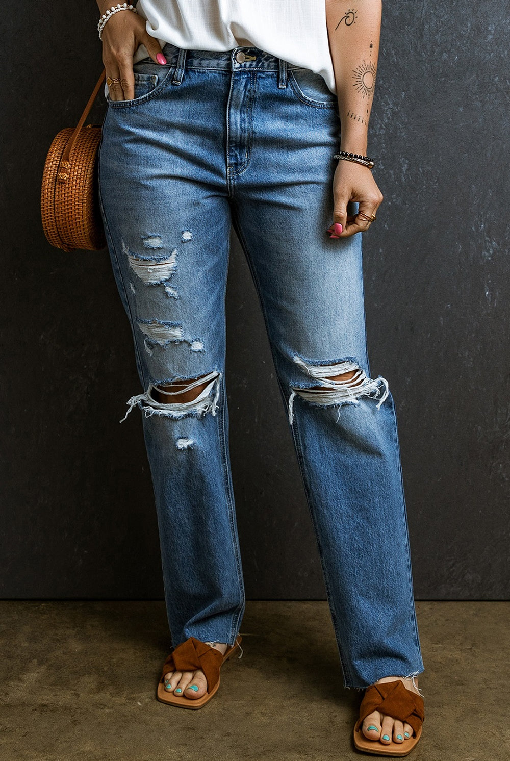 Close-up of distressed boyfriend jeans with strategic rips and a relaxed fit, paired with brown sandals and a woven bag.