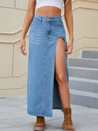 Woman in a light-wash high-waisted denim maxi skirt with a thigh-high slit, paired with a white crop top and brown lace-up boots.