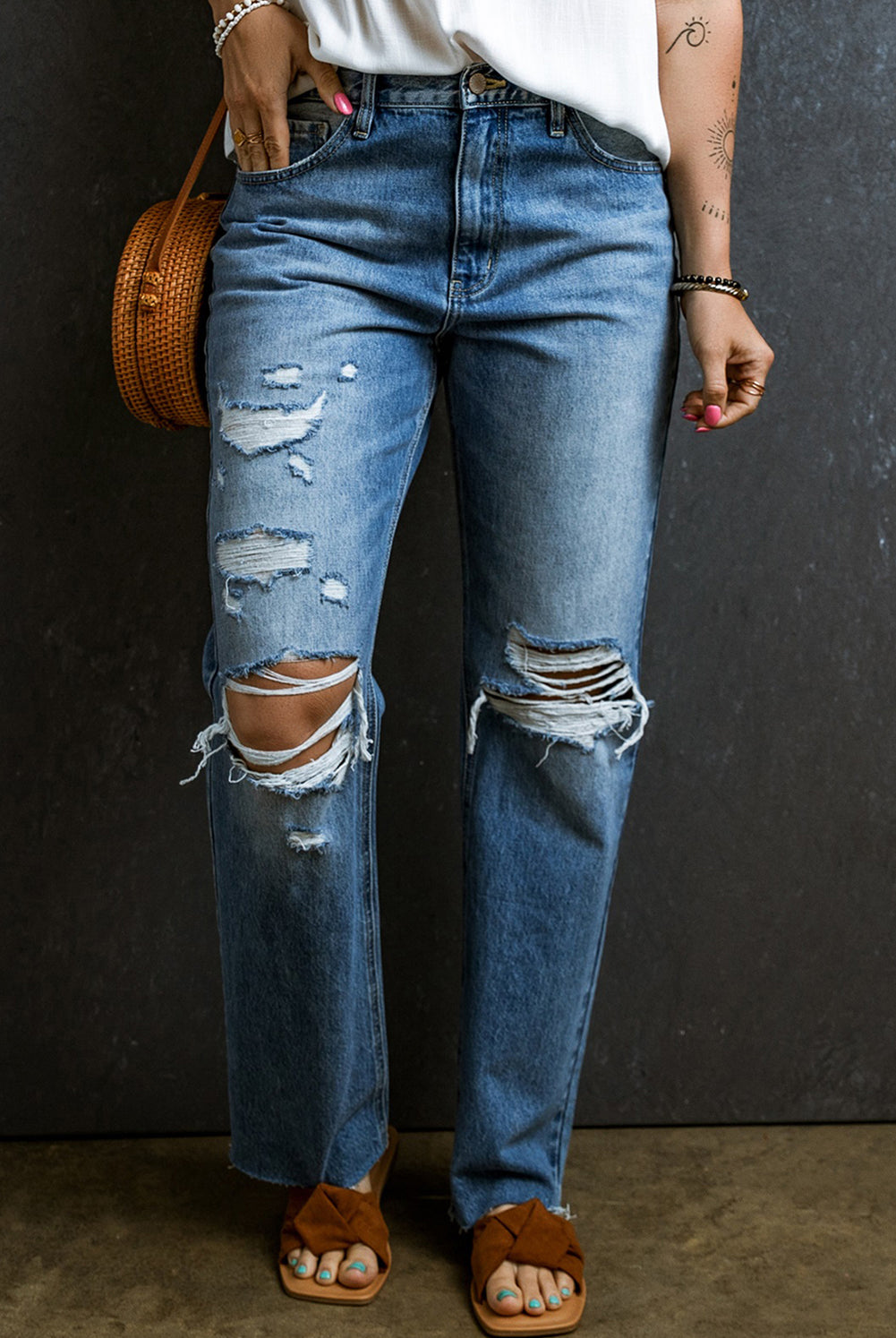 Close-up of distressed boyfriend jeans with strategic rips and a relaxed fit, paired with brown sandals and a woven bag.