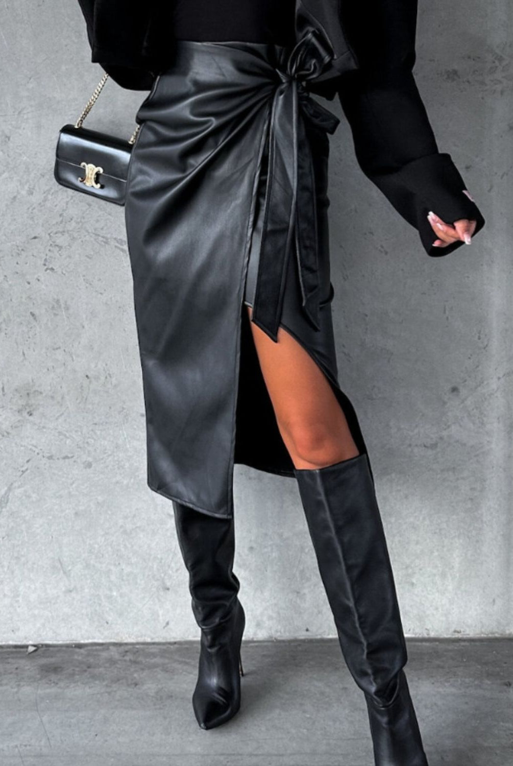 Woman posing in a chic high-waisted black skirt with tie detail and thigh-high slit, paired with a strapless top, knee-high boots, and a small black shoulder bag, against an industrial grey background.