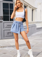 A model wearing a denim skort with a frayed hem, styled with a cropped white tank top, chunky white boots, and a black crossbody bag, creating a bold yet casual look.
