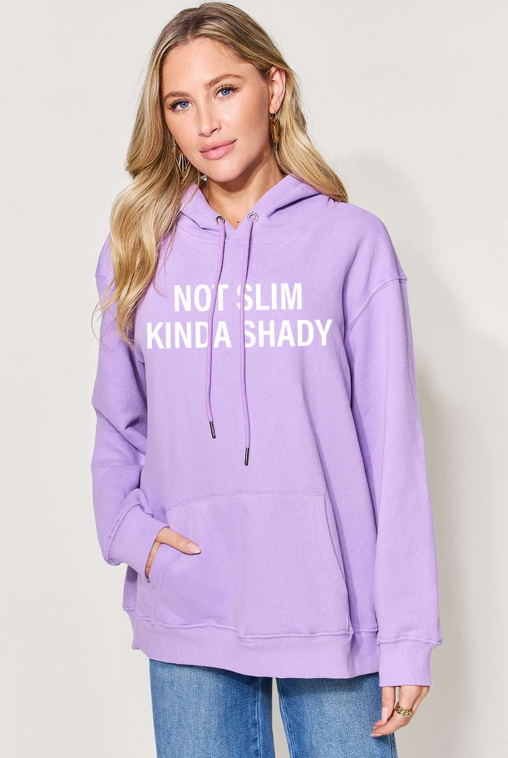 A woman wearing a black oversized hoodie with the text "NOT SLIM KINDA SHADY" printed in bold white letters across the front. The hoodie has a drawstring hood and a large kangaroo pocket. She is casually styled with light-wash denim jeans and has a natural makeup look with her blonde hair styled in loose waves. The overall vibe is relaxed and edgy.