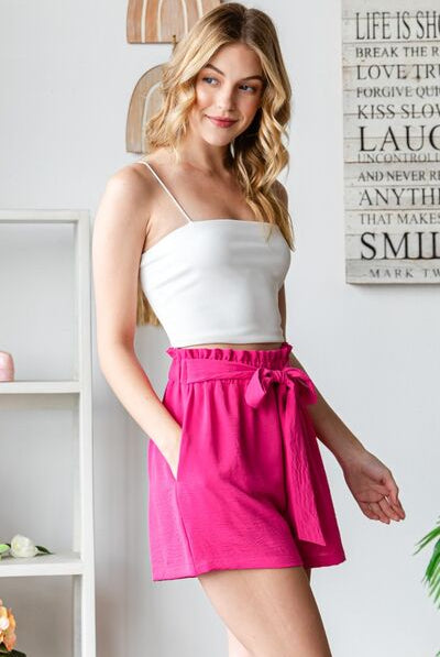 A model standing in a chic setting wearing GemThreads Boutique’s pink ruffle waist shorts paired with a white strapless top and beige ankle-strap heels.