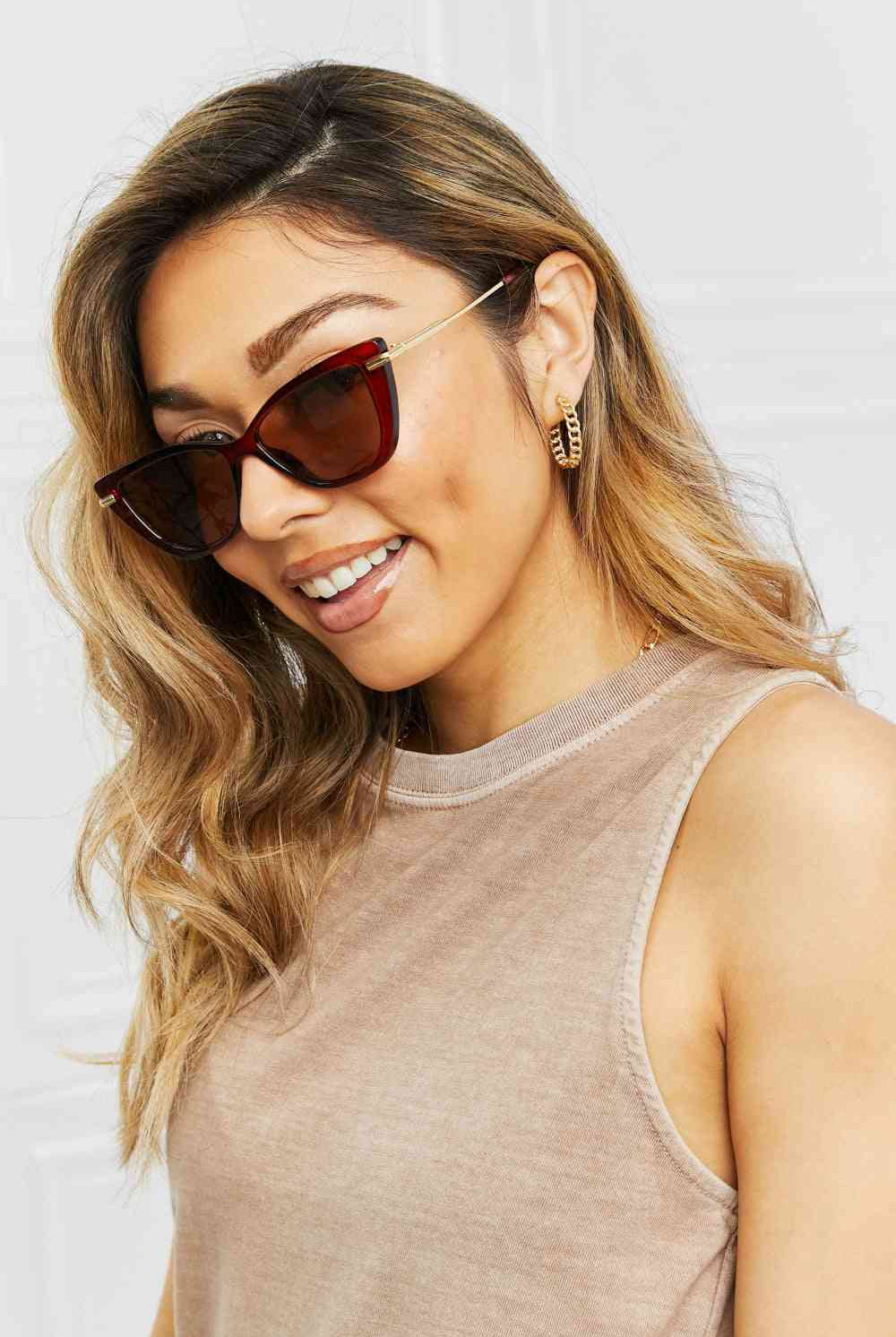 Woman wearing burgundy cat-eye sunglasses with a subtle gradient, paired with gold hoop earrings and a casual beige tank top, exuding a relaxed yet sophisticated summer vibe.