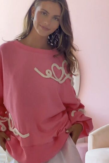 A model in a relaxed pose wearing a white round neck sweatshirt with pink 'XOXO' graphic text, paired with pink denim shorts, against a pink and white patterned background.