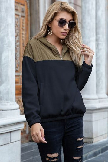 Woman in a stylish quarter-zip color-block hoodie and distressed jeans, accessorized with sunglasses and open-toe sandals, posing in an urban setting.