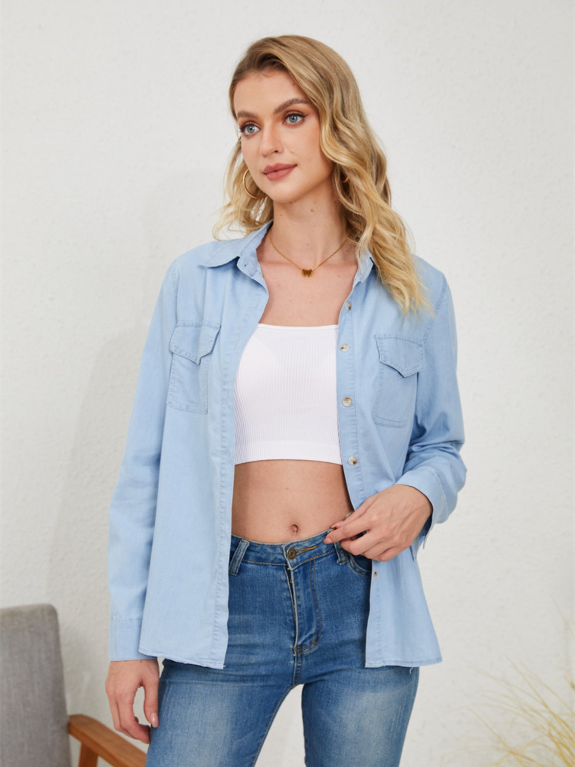 A woman is casually styled in a light denim button-up shirt, left open to reveal a white crop top underneath, and paired with blue jeans for a cohesive, double-denim look.