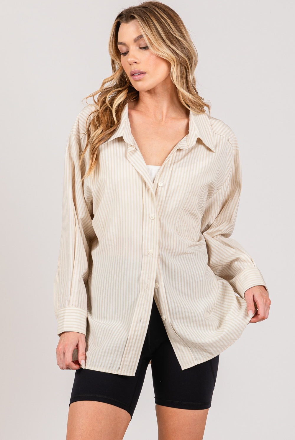 A woman is showcasing a chic beige striped long sleeve shirt with a casual open front design, layered over a white crop top, and paired with black biker shorts. The shirt features a classic collar, a chest pocket, and buttoned cuffs, highlighting a versatile and trendy piece from GemThreads Boutique.