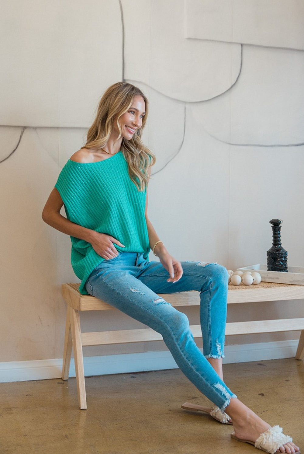 A woman smiling gently while modeling a teal ribbed sweater vest with an off-the-shoulder neckline, giving it a casual yet chic look. She is paired with light blue distressed jeans, sitting on a wooden stool against a neutral background, adding a relaxed and approachable vibe to the image. Her blonde hair falls in loose waves, complementing the laid-back yet fashionable outfit.
