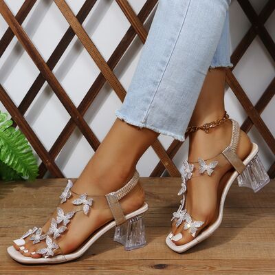 Elegant heeled sandals adorned with sparkling butterfly embellishments and a clear block heel, paired with chic ankle bracelets and frayed blue jeans.
