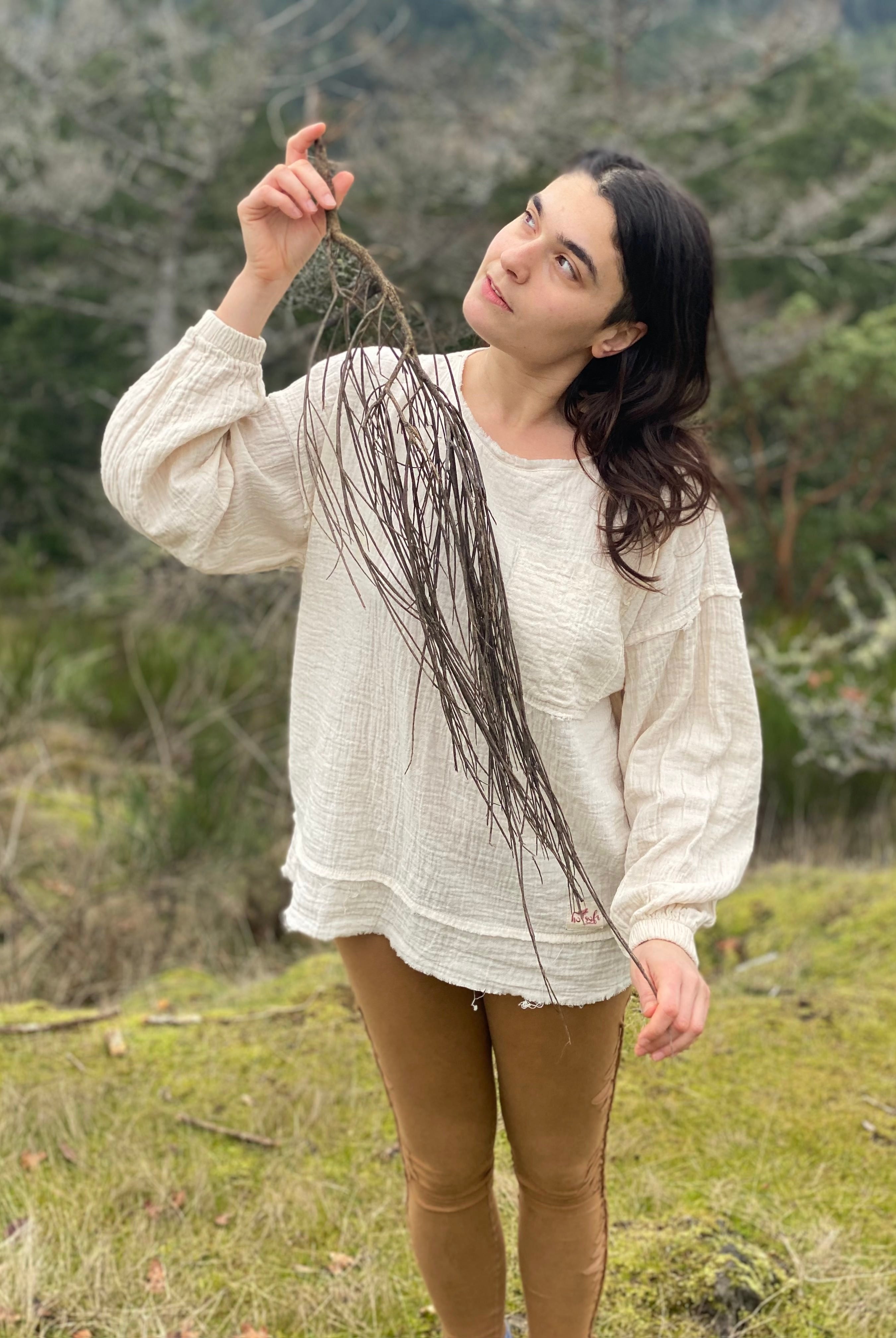 This image shows a woman in a natural outdoor setting, enjoying a moment of tranquility. She is dressed in an off-white, textured, long-sleeve blouse with a relaxed fit, paired with form-fitting, olive-green leggings and appears to be holding a thin branch or twig, eyes closed, seemingly appreciating the scent or texture of nature's offering.
