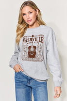 A woman models a heather gray 'Nashville Music City Tennessee' sweatshirt, displaying the vintage print design on the front, paired with blue jeans for a relaxed look.