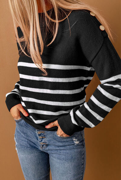 Smiling woman wearing a brown sweater with black horizontal stripes and button details on the shoulder, paired with white ripped jeans, accessorized with hoop earrings and a necklace.