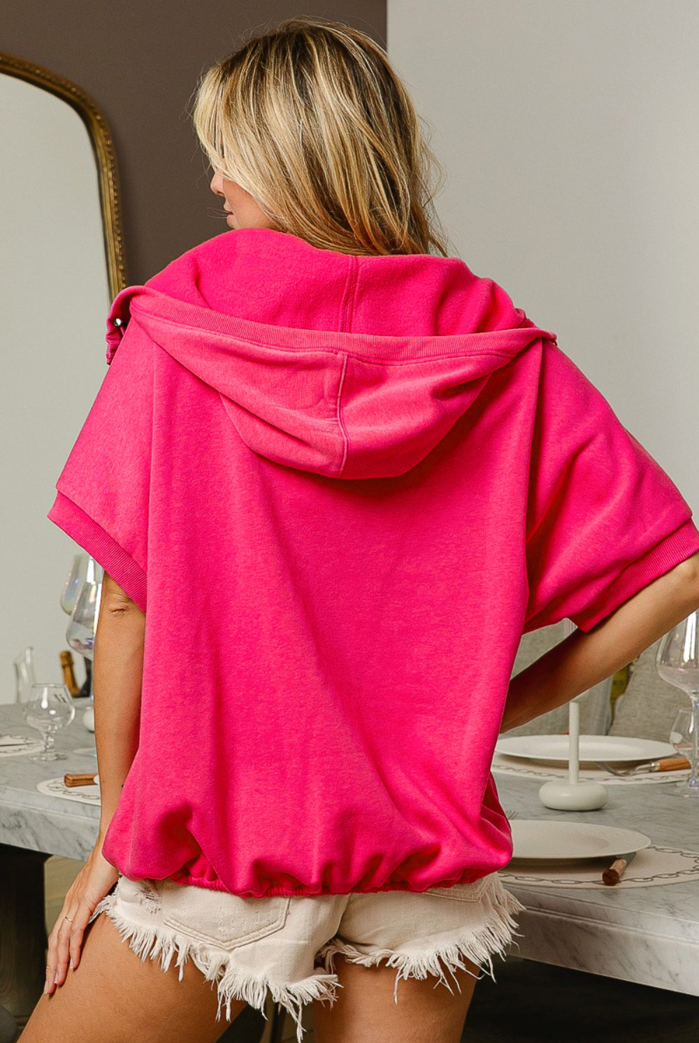 A woman models a vibrant pink short sleeve half zip up French Terry hoodie, with the hood draped elegantly over her wavy blonde hair. She stands in a stylish room, looking off to the side, with the zipper slightly open to give a relaxed vibe. The hoodie features a kangaroo pocket, adding a casual touch to the chic and sporty look.