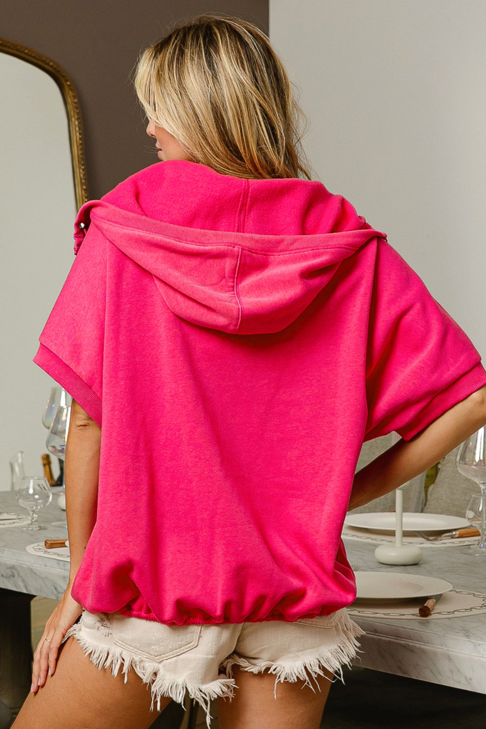 A woman models a vibrant pink short sleeve half zip up French Terry hoodie, with the hood draped elegantly over her wavy blonde hair. She stands in a stylish room, looking off to the side, with the zipper slightly open to give a relaxed vibe. The hoodie features a kangaroo pocket, adding a casual touch to the chic and sporty look.