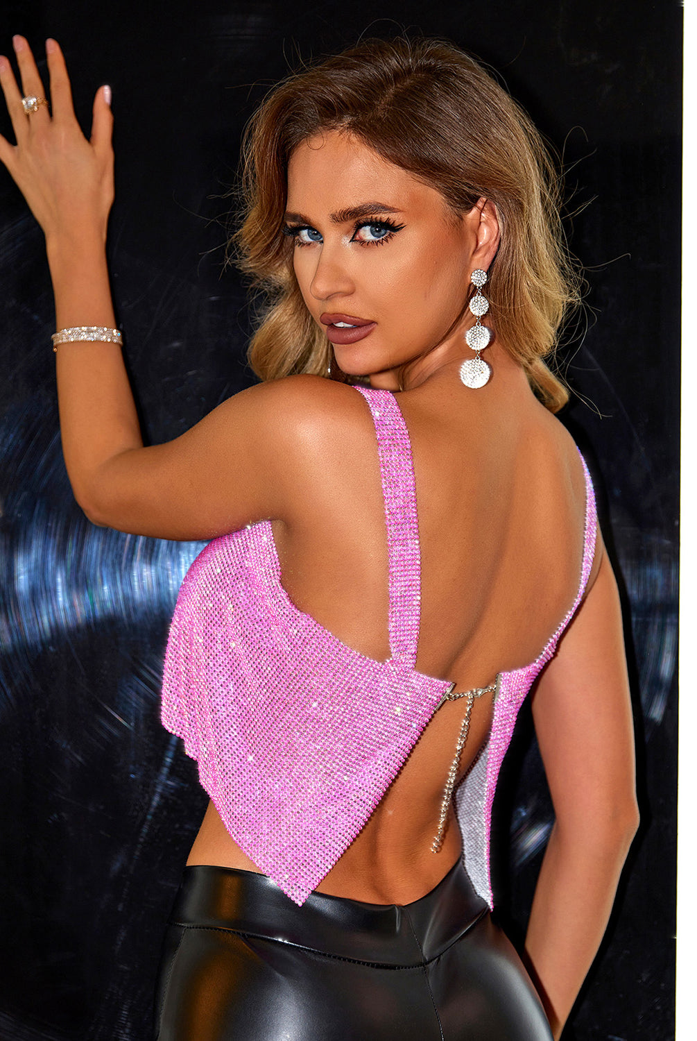 A woman modeling a shimmering pink sequin crop top with wide shoulder straps and a square neckline. The top is paired with high-waisted black faux leather pants, accentuating a trendy and sophisticated look. She complements the outfit with sparkling teardrop earrings and a matching silver bracelet, creating a cohesive and glamorous ensemble. Her hair is styled in elegant waves, and her makeup features a bold eye and a soft, matte lip color, highlighting her confident and stylish appearance.