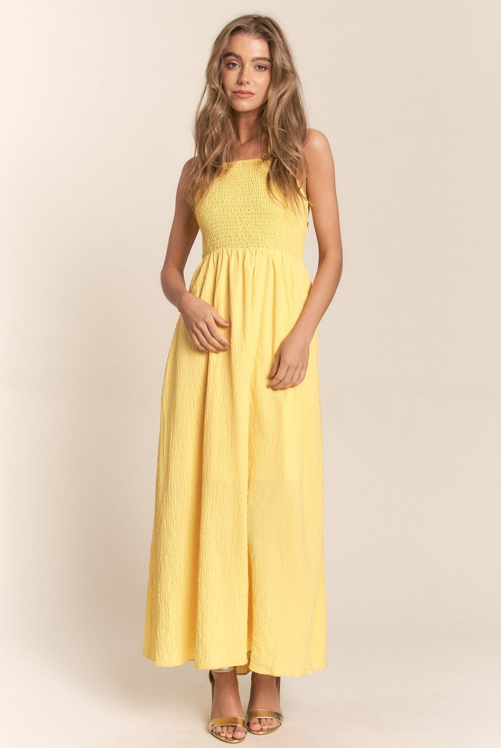 A woman glances over her shoulder, wearing a sunny yellow maxi dress with a unique ruffle detail running asymmetrically across the back. The dress ties at the lower back, creating an elegant bow that enhances the feminine silhouette. Its crinkled texture adds a bohemian flair to the design, perfect for summer days or a tropical getaway.