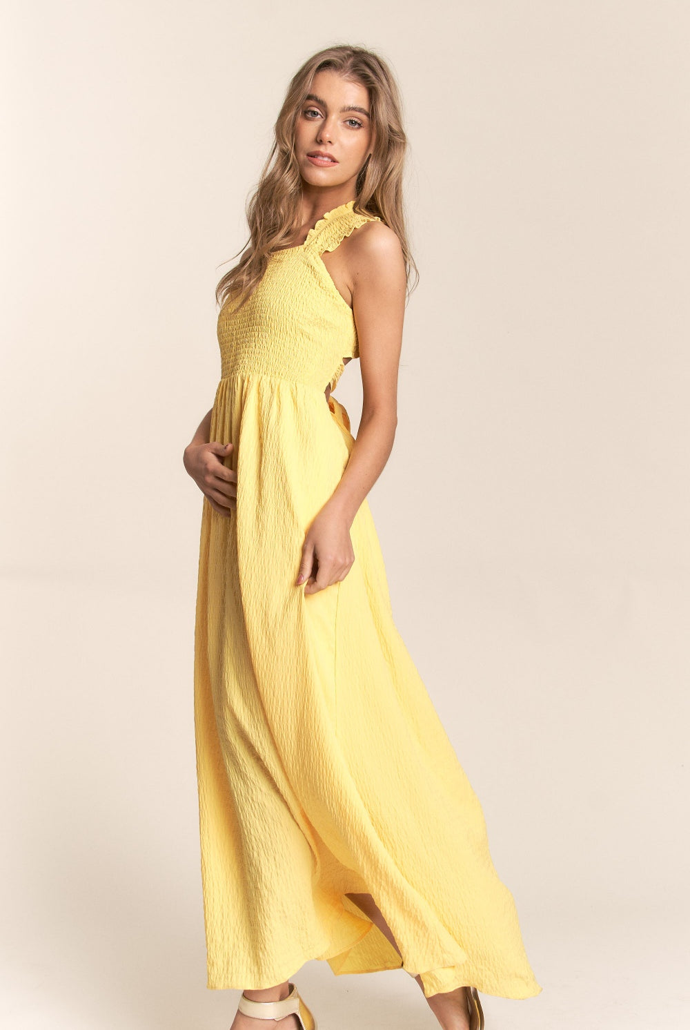 A woman glances over her shoulder, wearing a sunny yellow maxi dress with a unique ruffle detail running asymmetrically across the back. The dress ties at the lower back, creating an elegant bow that enhances the feminine silhouette. Its crinkled texture adds a bohemian flair to the design, perfect for summer days or a tropical getaway.