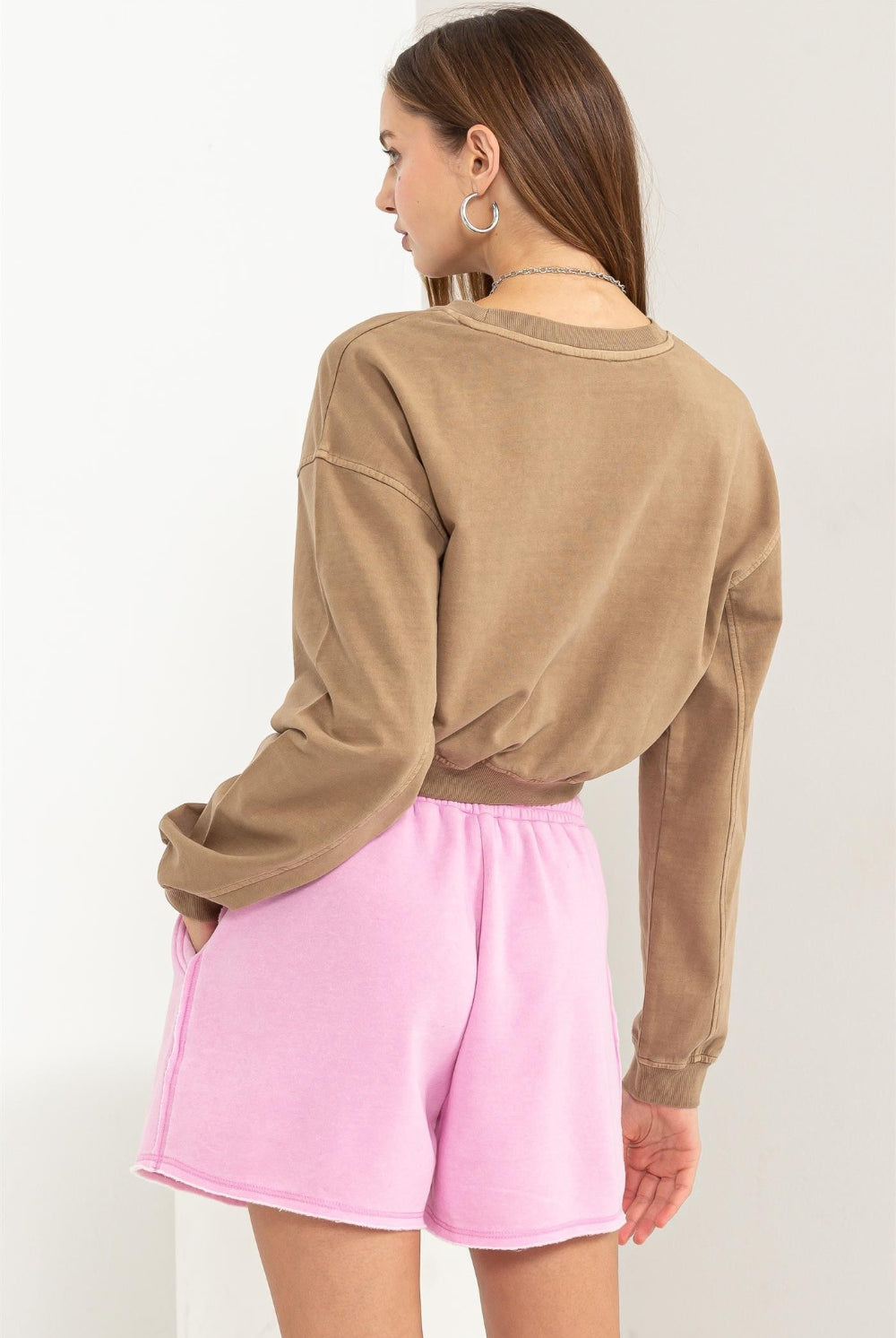 A woman modeling a HYFVE round neck long sleeve cropped sweatshirt in a solid khaki color. The sweatshirt has a raw hemline and drop shoulders, giving off a relaxed and trendy vibe. She is also wearing pink drawstring shorts, complementing the casual look.