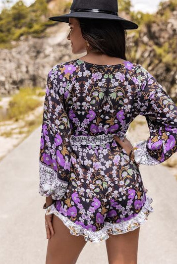 Stylish woman in a purple floral romper with lace trim, wide sleeves, and ruffled hem, paired with black cowboy boots and a wide-brimmed hat.