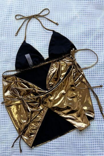 Model posing in a luxurious gold halter neck three-piece bikini set, featuring tie-side bottoms and a coordinating sarong, against a textured backdrop.