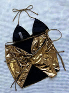 Model posing in a luxurious gold halter neck three-piece bikini set, featuring tie-side bottoms and a coordinating sarong, against a textured backdrop.