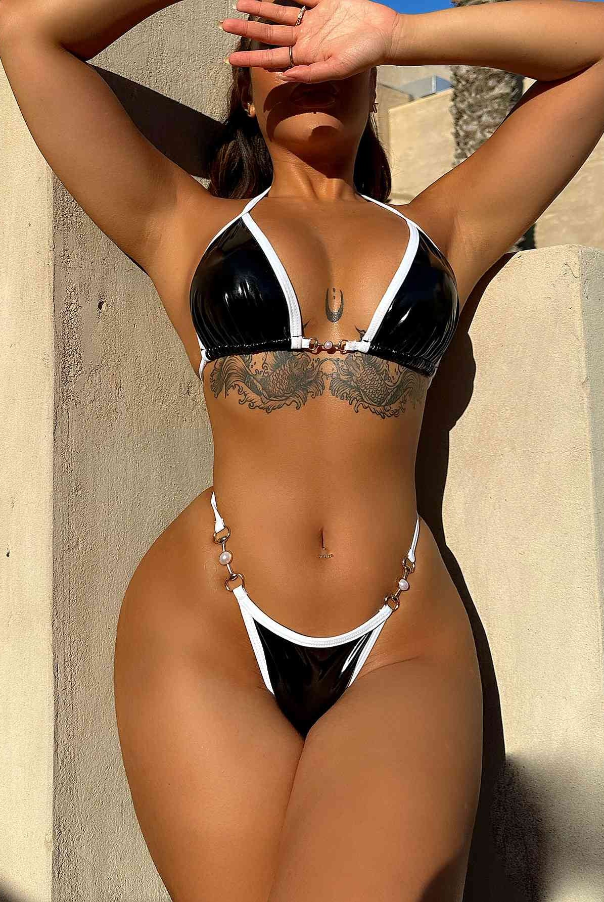 Woman posing in a black and white halter neck bikini set with metallic accents and ring details, against a warm, sunlit wall.
