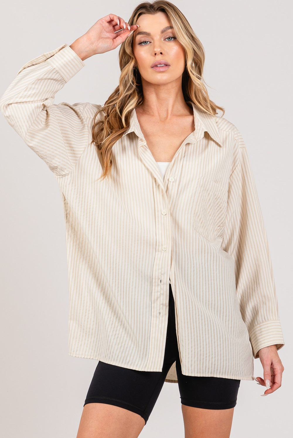 A woman is showcasing a chic beige striped long sleeve shirt with a casual open front design, layered over a white crop top, and paired with black biker shorts. The shirt features a classic collar, a chest pocket, and buttoned cuffs, highlighting a versatile and trendy piece from GemThreads Boutique.