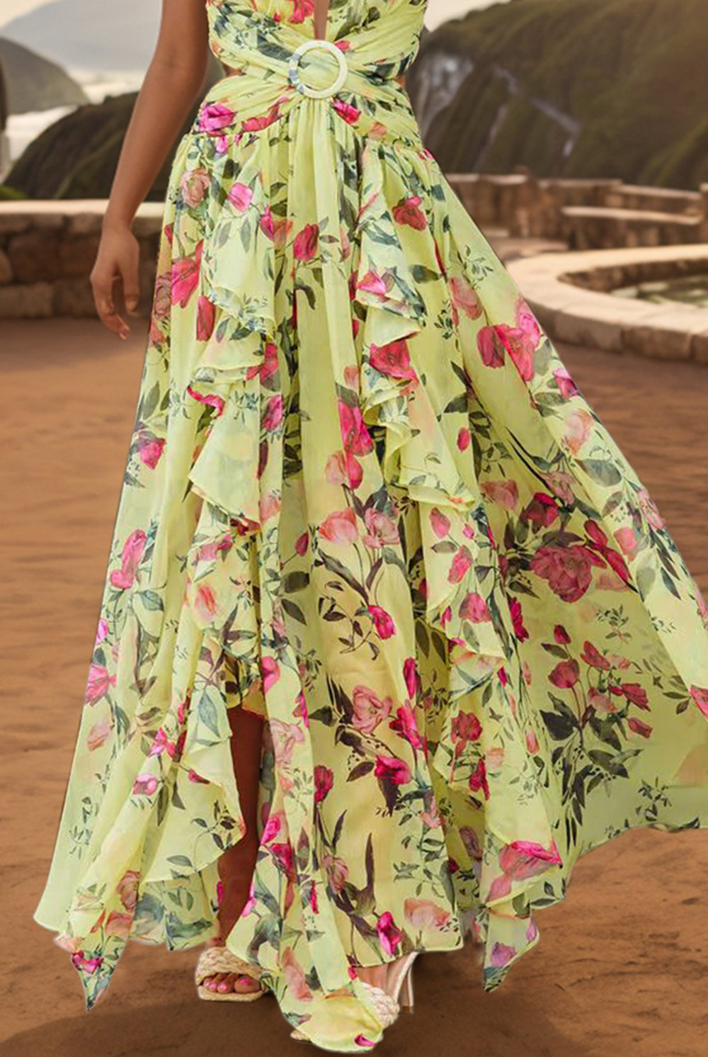 A woman stands poised against a sunset backdrop, wearing an elegant sleeveless maxi dress adorned with a vibrant floral pattern in shades of pink and green. The dress features a plunging neckline, cinched waist with a decorative ring detail, and a flowing ruffled skirt, embodying a graceful summer evening aesthetic.
