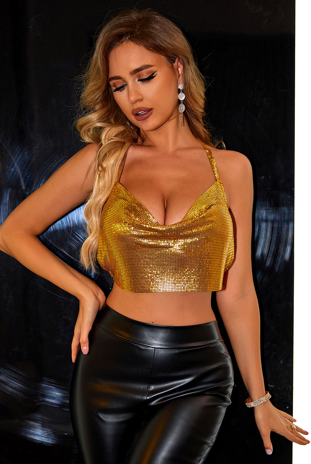 A model poses in a striking gold sequin cami top with a cowl neckline and thin straps, paired with high-waisted black leather pants. The top shimmers with every movement, exuding luxury and glamour, while the model's accessorizing with elegant teardrop earrings and a sparkling bracelet completes the sophisticated look. Her makeup is dramatic and polished, with smoky eyes and a matte lip, and her wavy blonde hair cascades over one shoulder, emphasizing the opulence of the ensemble.