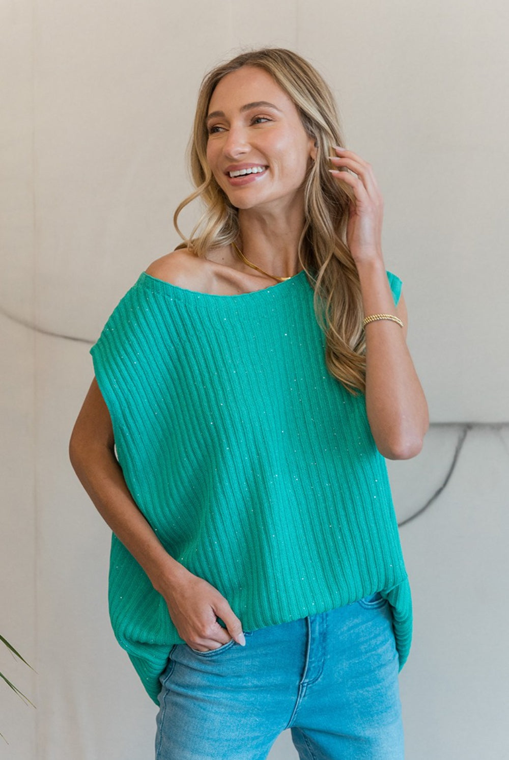 A woman smiling gently while modeling a teal ribbed sweater vest with an off-the-shoulder neckline, giving it a casual yet chic look. She is paired with light blue distressed jeans, sitting on a wooden stool against a neutral background, adding a relaxed and approachable vibe to the image. Her blonde hair falls in loose waves, complementing the laid-back yet fashionable outfit.