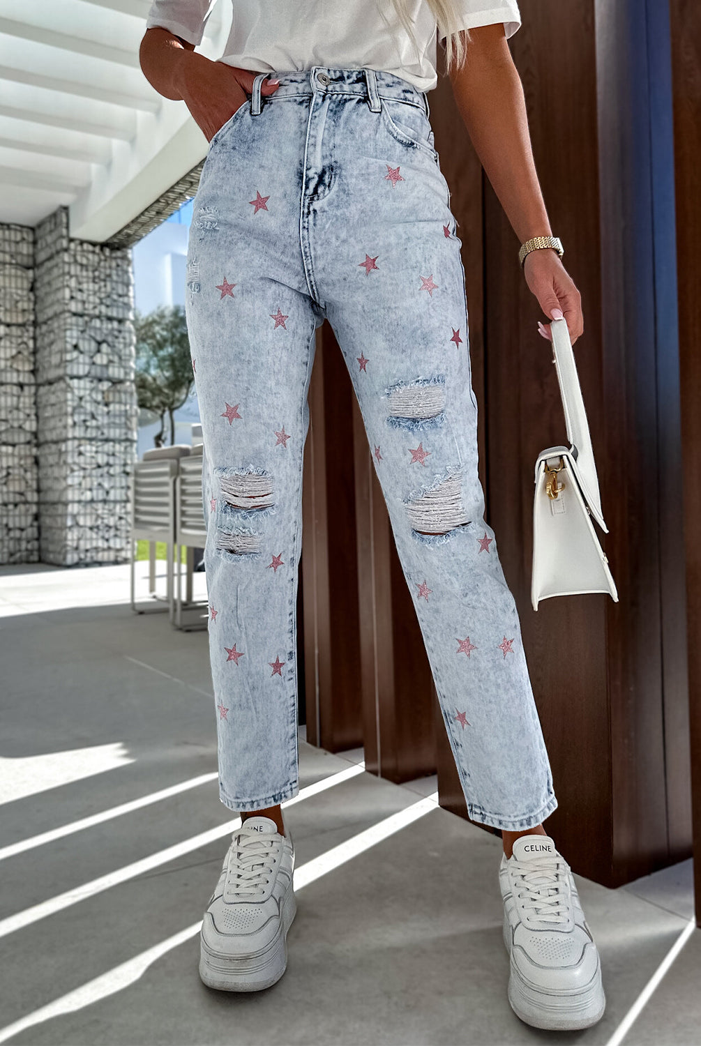 Fashion-forward woman sporting distressed light-wash high-waist jeans with a unique red star pattern, paired with classic white sneakers for a chic, street-style look.
