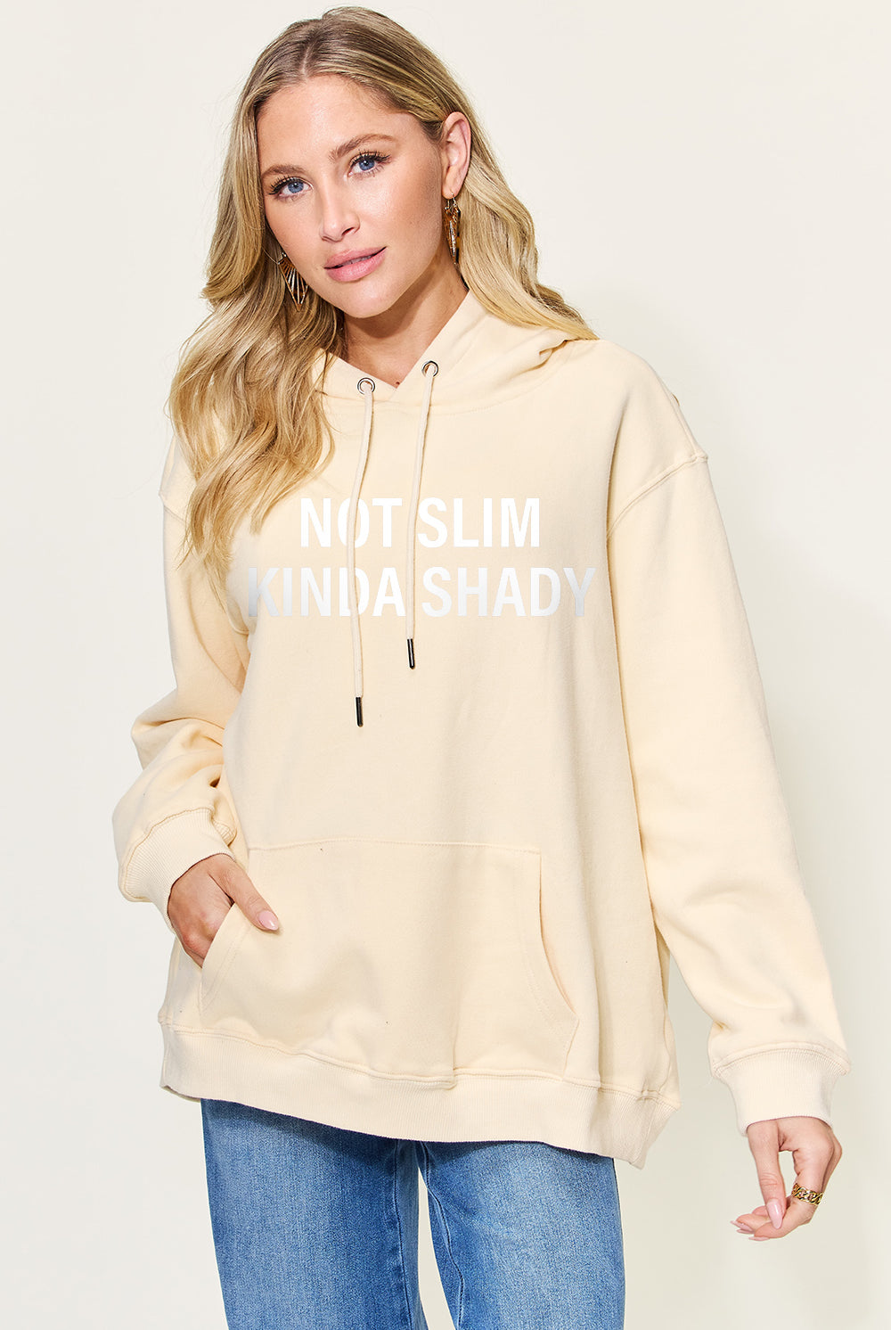 A woman wearing a black oversized hoodie with the text "NOT SLIM KINDA SHADY" printed in bold white letters across the front. The hoodie has a drawstring hood and a large kangaroo pocket. She is casually styled with light-wash denim jeans and has a natural makeup look with her blonde hair styled in loose waves. The overall vibe is relaxed and edgy.