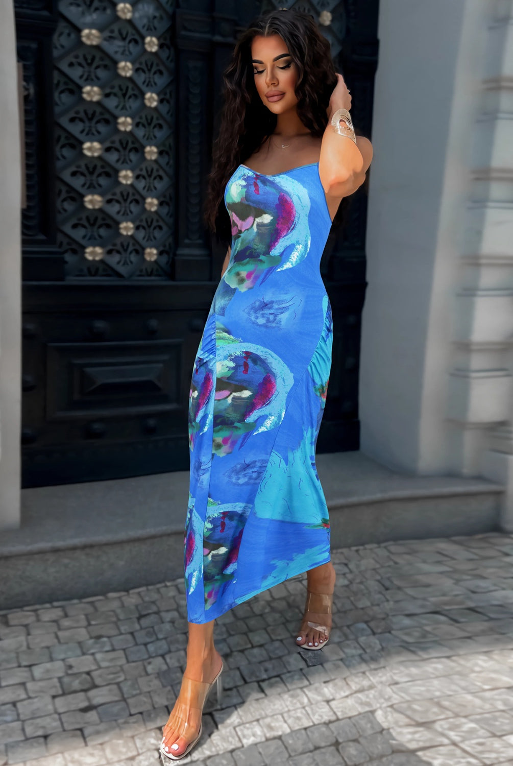 Woman wearing a printed spaghetti strap cami dress, paired with clear heels and holding a white bag.