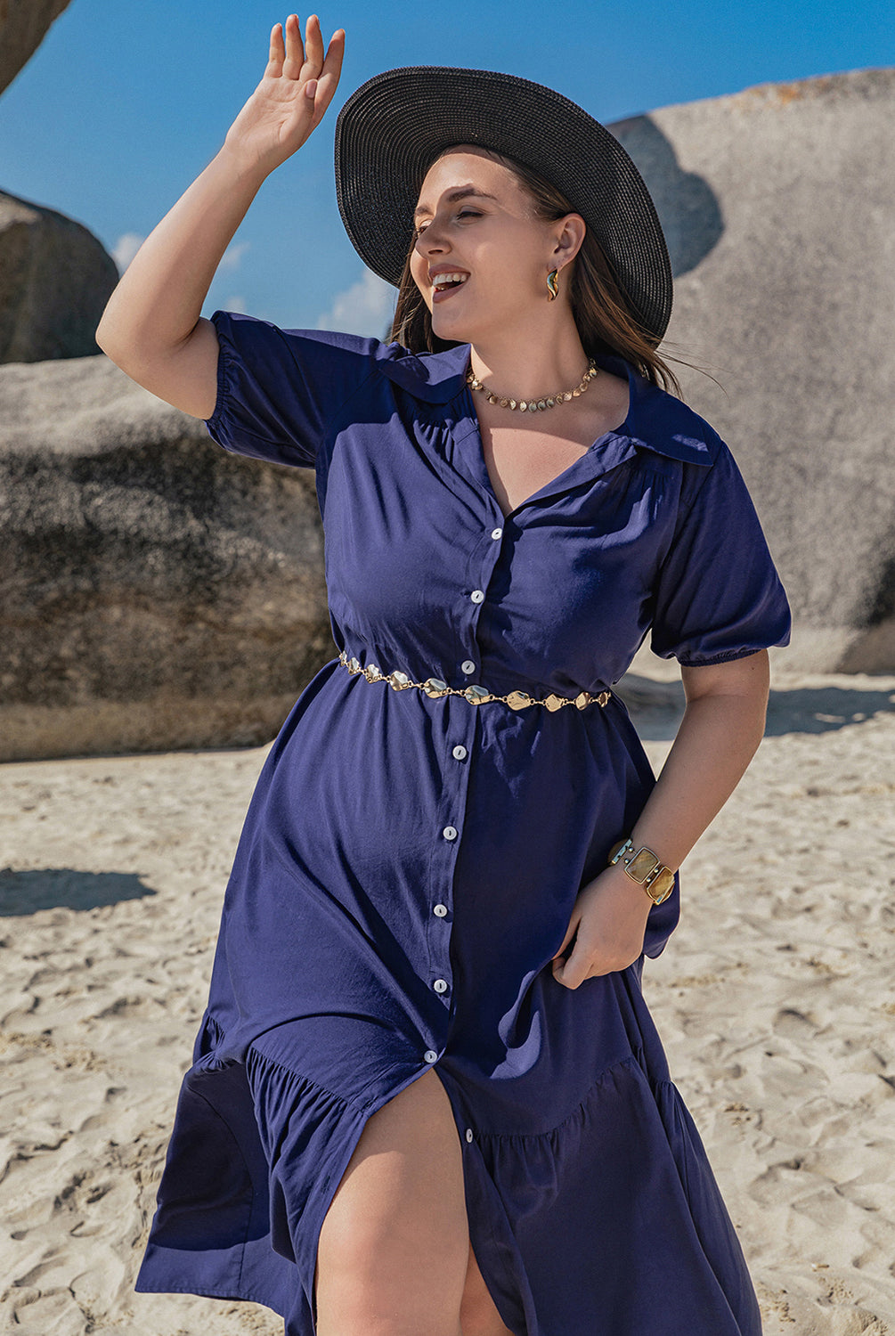 Woman wearing a plus size blue collared short sleeve midi dress, standing on a sandy beach with a large rock formation in the background.