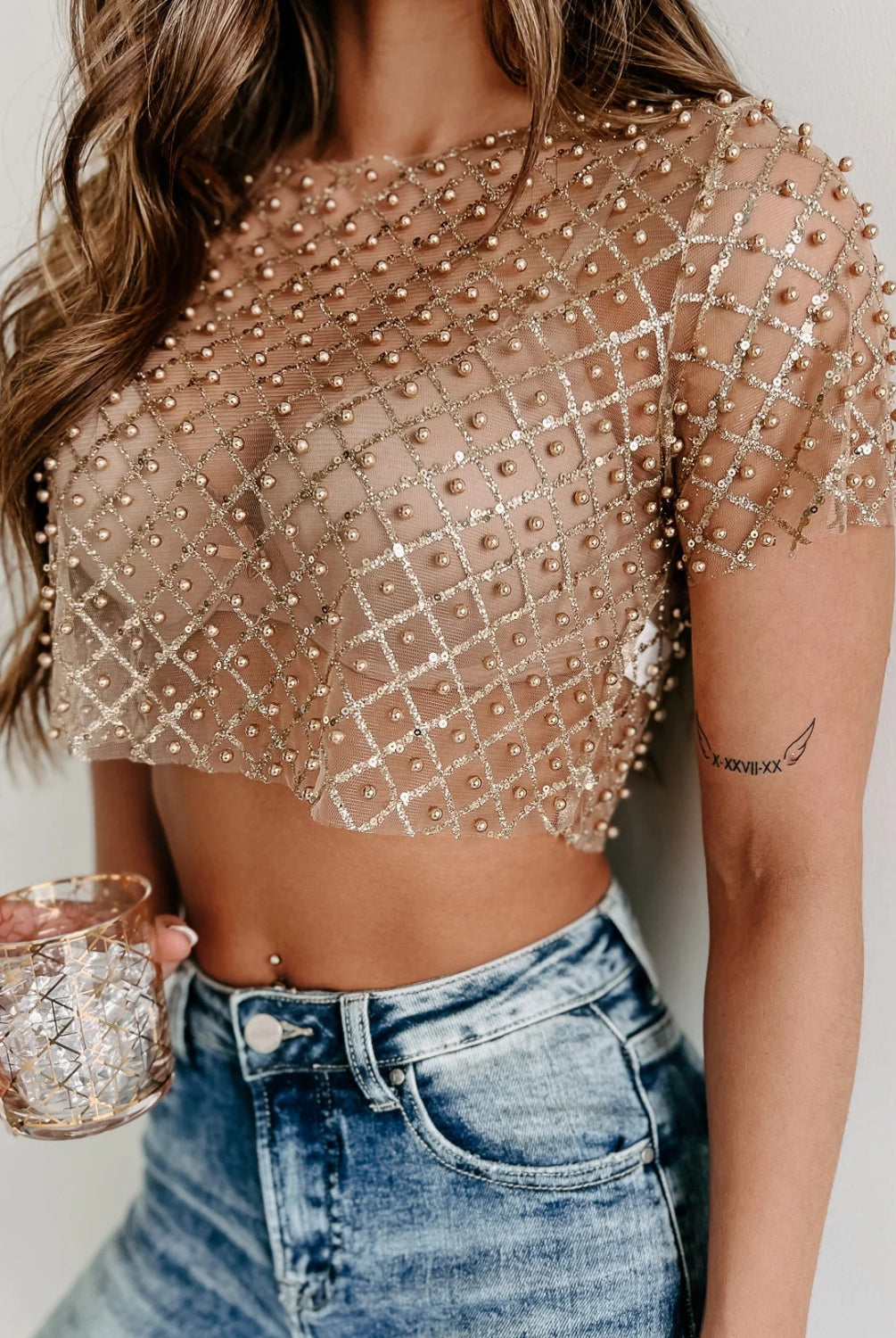 A close-up of a woman wearing a sheer, short-sleeved blouse embellished with beads and sequins in a grid pattern, paired with high-waisted blue jeans. She's holding a glass in one hand and a cream-colored chain strap purse in the other. The blouse is a glamorous, cropped design that reveals the midriff.