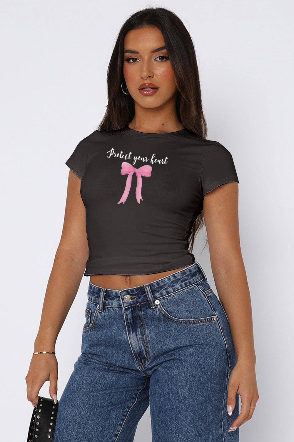 A woman wearing a fitted, crew-neck graphic t-shirt with the phrase "Protect your heart" above a ribbon design, paired with casual denim jeans and accessorized with chic sunglasses and simple jewelry. The shirt has a cropped cut, offering a modern and stylish look.