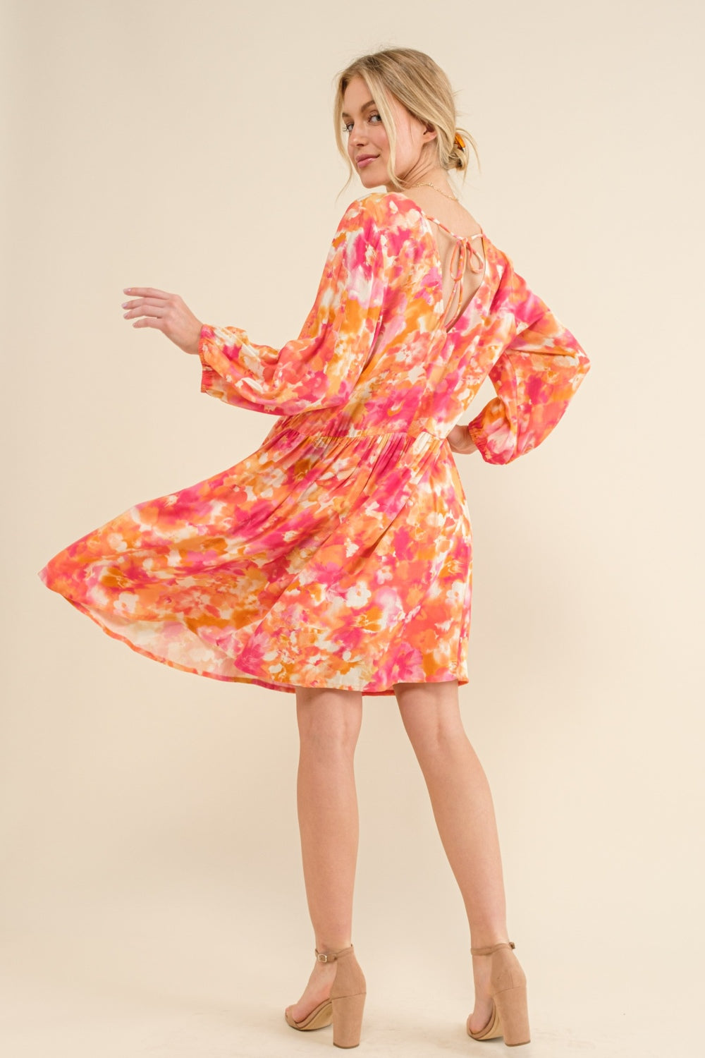 Woman wearing a vibrant floral print long sleeve dress in shades of orange and pink, featuring a tie back detail.