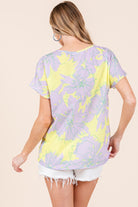 A woman wearing a vibrant yellow and purple floral short sleeve t-shirt, styled casually with white distressed shorts for a fresh, summery look.