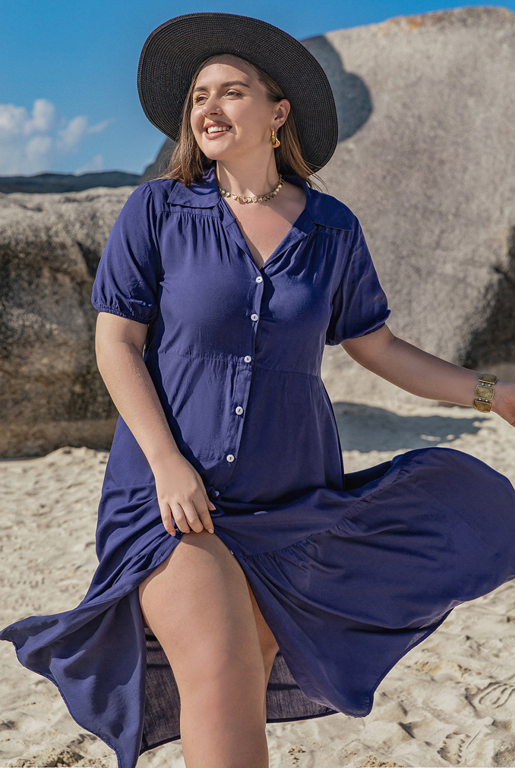Woman wearing a plus size blue collared short sleeve midi dress, standing on a sandy beach with a large rock formation in the background.