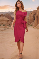 Woman wearing a magenta one-shoulder wrap dress with side tie detail, standing in a desert landscape.