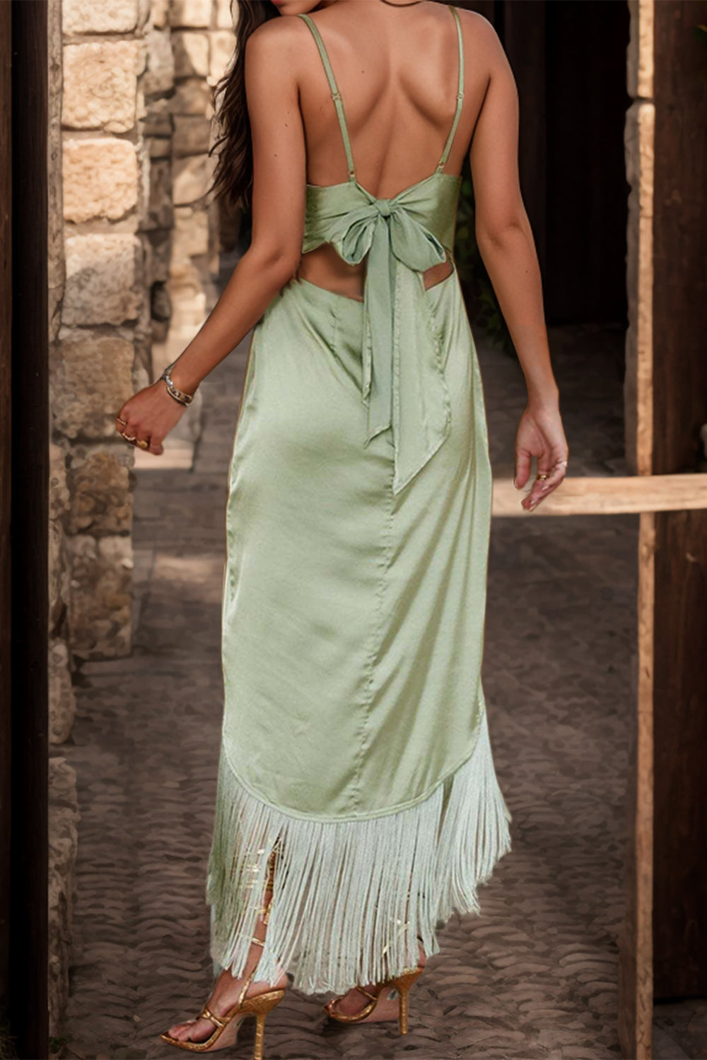 A model in a cami dress with a sleek bodice, adjustable straps, and an asymmetrical hemline enhanced by a fringe of tassels, paired with elegant strap sandals, exuding a playful yet sophisticated aura.